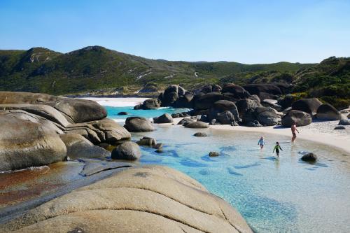 large granite boulders on beach surrounded by clear blue water and a person and children playing in the water 