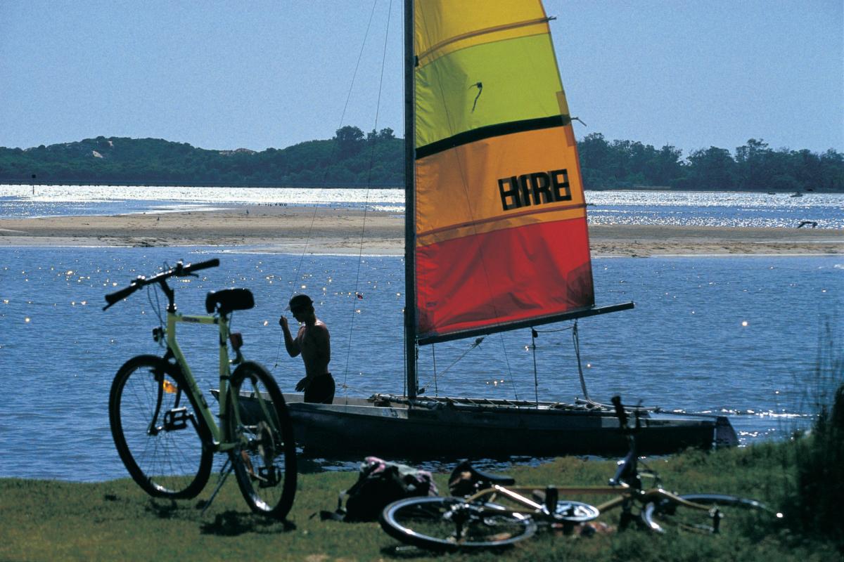 man stabnding in water near catamaran with orange and red sail and two bicycles nearby on grass