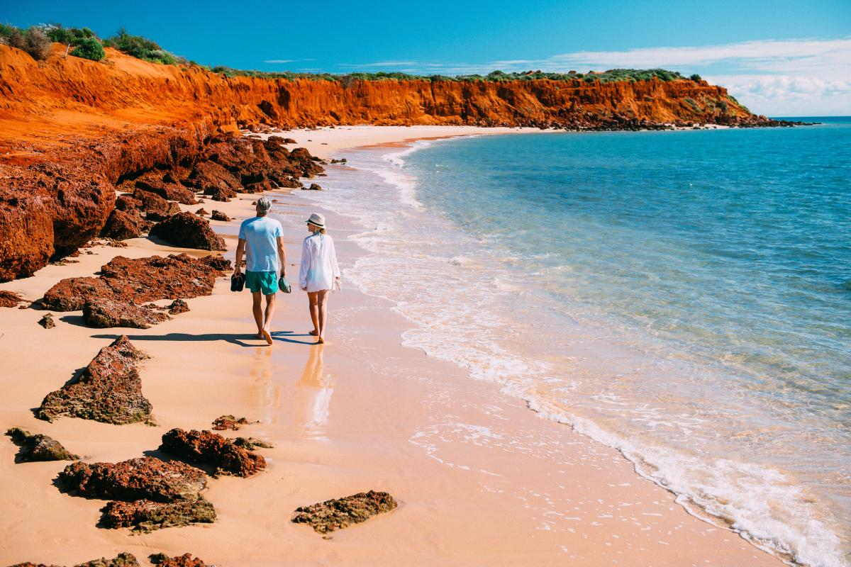 Visitors walking along beach on colourful coastal bay of red dirt, white sand and calm green blue ocean.
